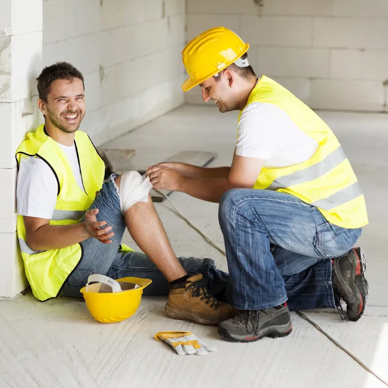 A construction worker with a hard hat and safety vest sits on the floor, smiling, despite a knee injury. Another worker in similar attire kneels beside him, tending to the wound. The scene is set indoors amidst ongoing construction, highlighting the need for efficient doctor management services in such environments.