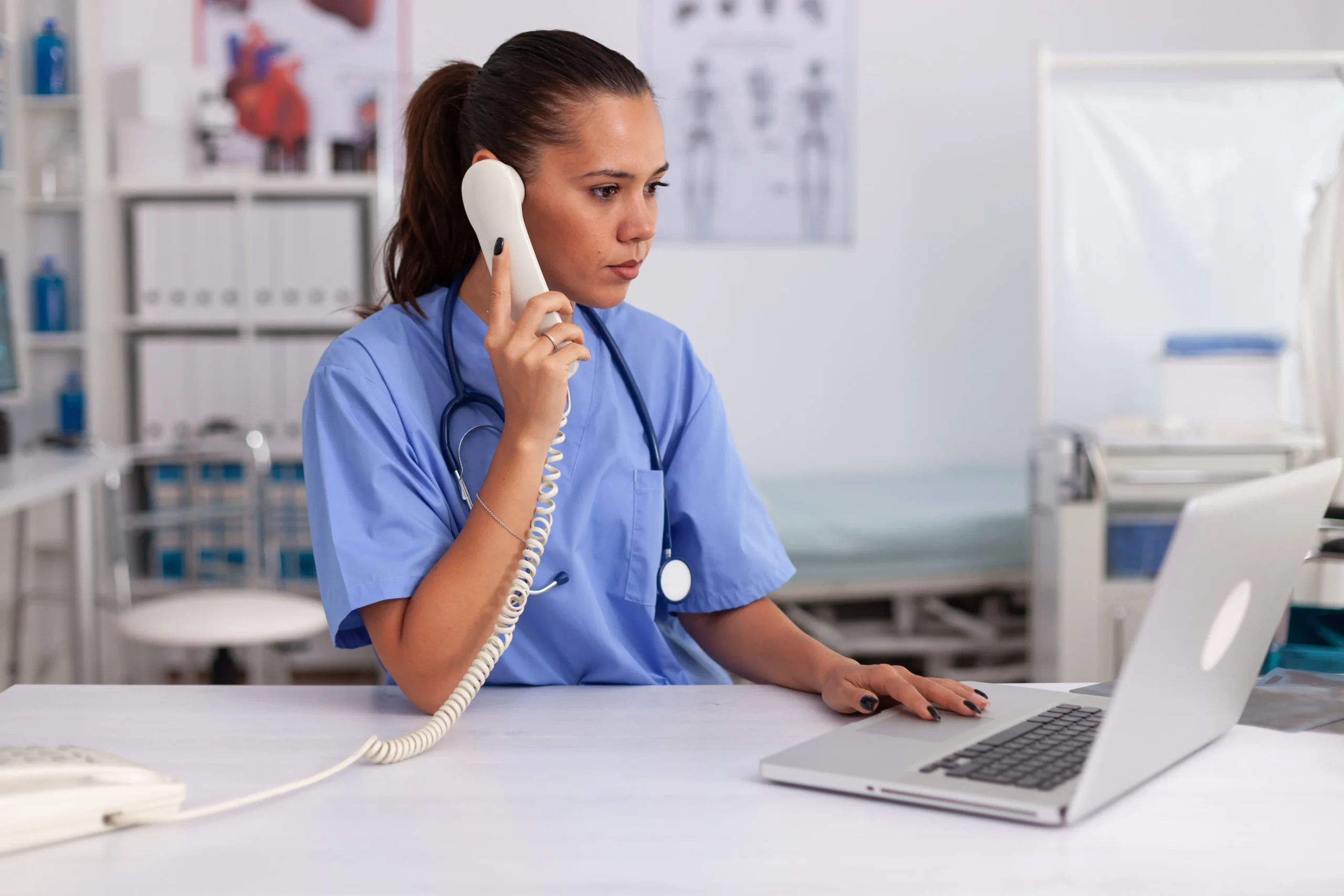 A medical professional wearing blue scrubs and a stethoscope around her neck is sitting at a desk, holding a telephone receiver to her ear, and looking at a laptop. The background includes medical equipment and charts on the wall, likely related to Doctor MGT services.