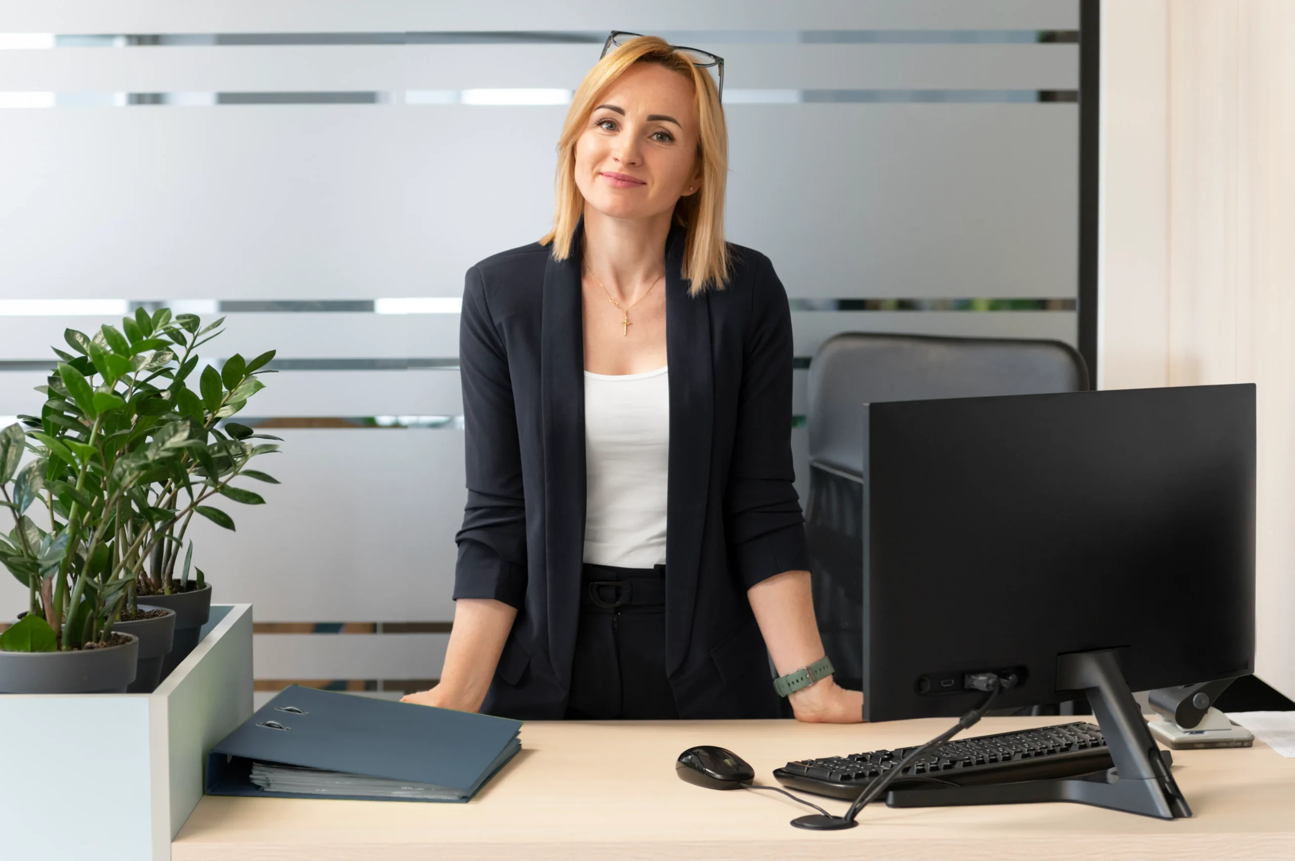 A woman with blonde hair and glasses on her head is standing behind a desk and smiling. She is dressed in a black blazer and white top. The desk has a computer monitor, keyboard, mouse, and some potted plants. The background features frosted glass panels with the logo of Doctor MGT.