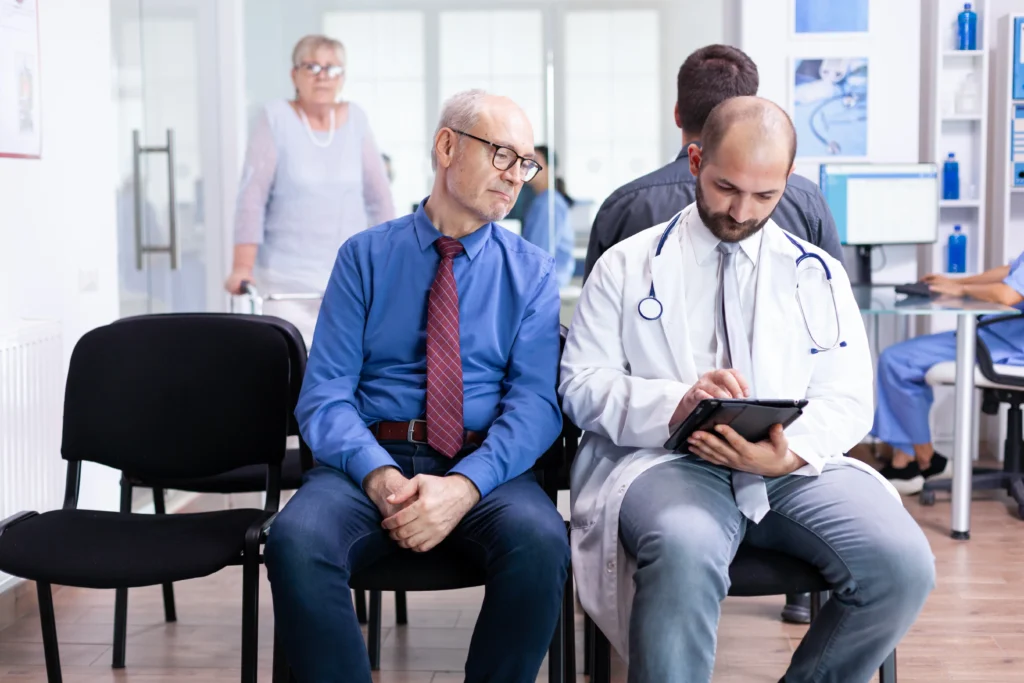 An older man with glasses and a tie sits beside a young doctor who is holding a tablet in a waiting area. The doctor, likely involved in Doctor MGT services, is focused on the tablet, while a woman with a walker is in the background. Other medical staff and patients can be seen further back.