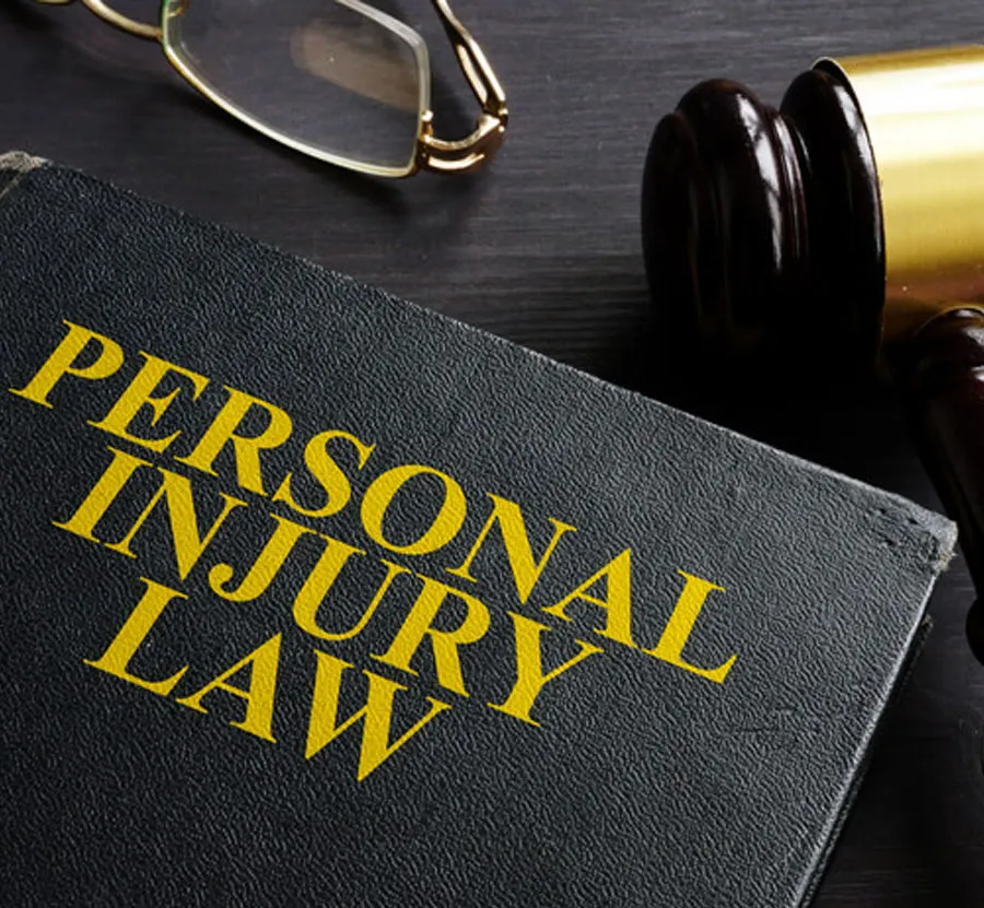 A book titled "PERSONAL INJURY LAW" with bold yellow letters sits on a dark wooden surface. Beside it are a pair of eyeglasses, a wooden judge's gavel, and some notes on medical billing and coding.