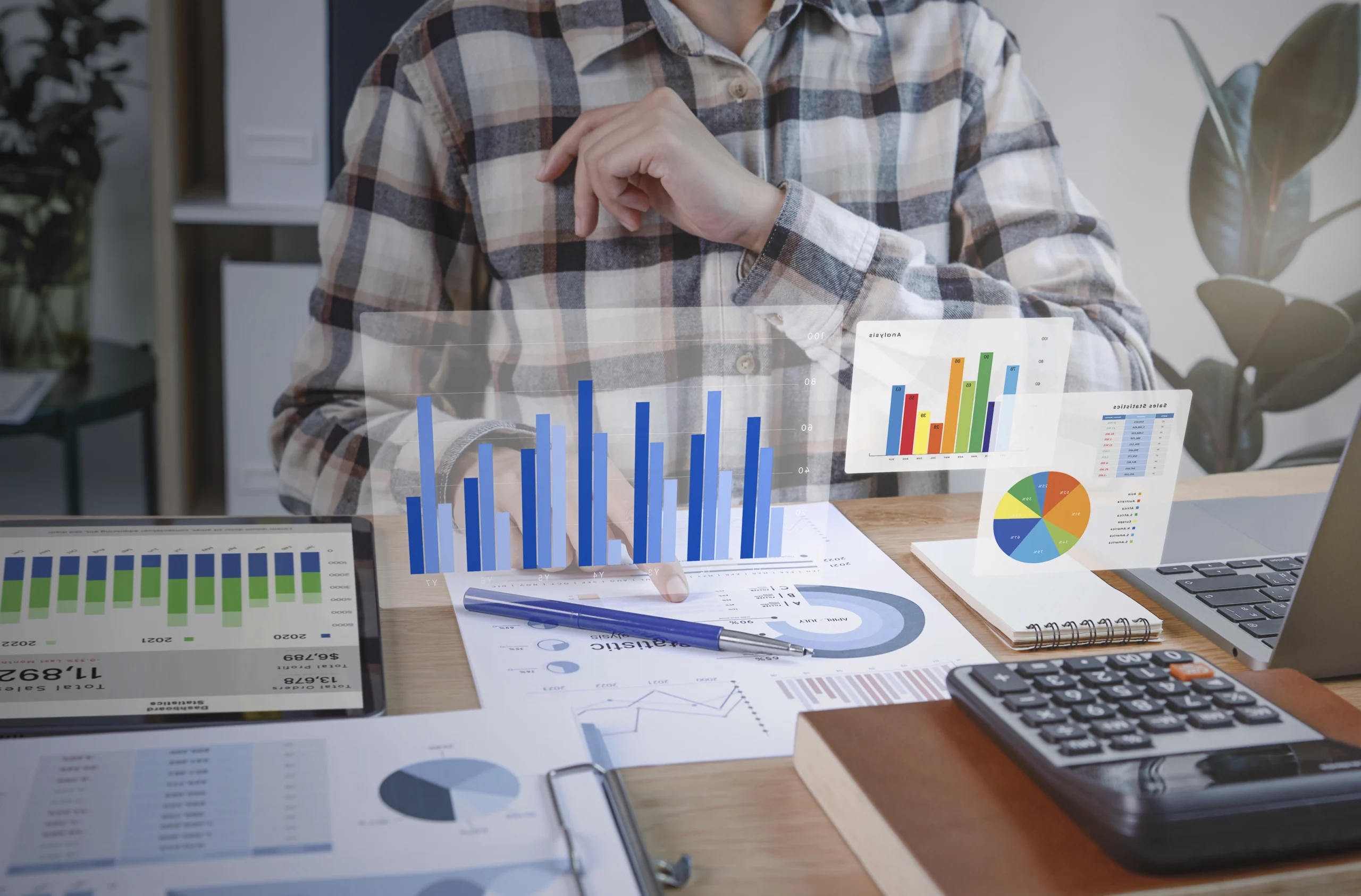 A person sits at a desk with various charts and graphs, both on paper and on transparent screens. They are surrounded by documents, a tablet, a calculator, and a notebook. The individual appears to be analyzing data and statistics in an office setting for Doctor management services.