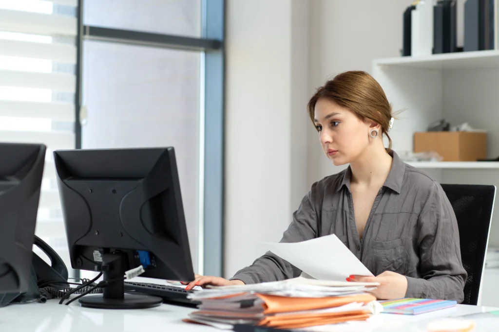A woman with light brown hair is sitting at a desk in a modern office, working on a computer. She is holding some papers and has multiple files and folders on the desk in front of her, related to medical billing and coding. There are shelves with office supplies in the background.