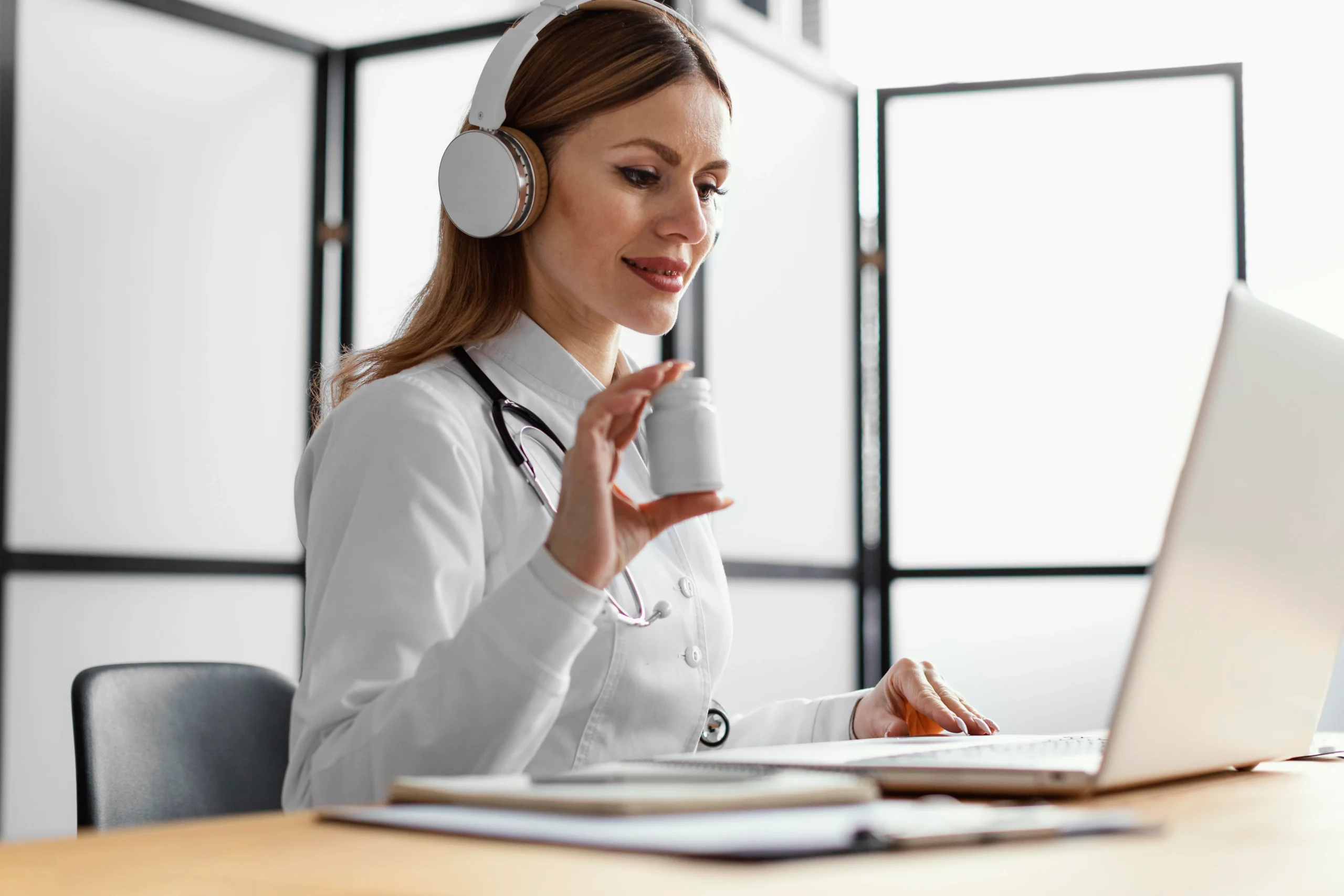 A female doctor with long brown hair, wearing a white coat and headphones, sits at a table with a laptop. She holds up a small white bottle while looking at the screen, possibly during a virtual consultation for Doctor MGT services. A stethoscope hangs around her neck.