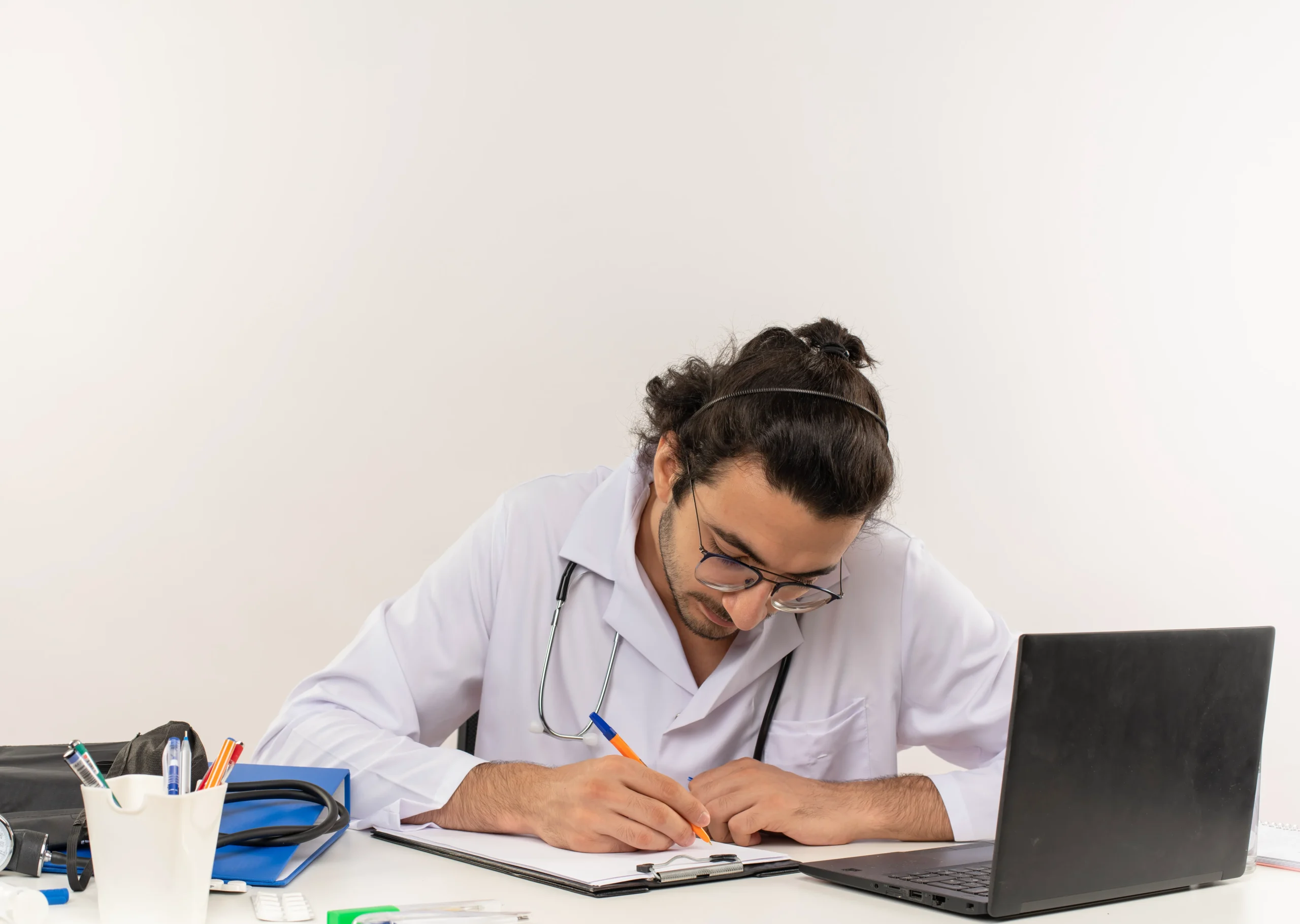 A doctor with a stethoscope around their neck is sitting at a desk, writing in a notebook with a pen. An open laptop and a cup holding various pens sit nearby, along with other items essential for Doctor MGT services. The background is plain white.