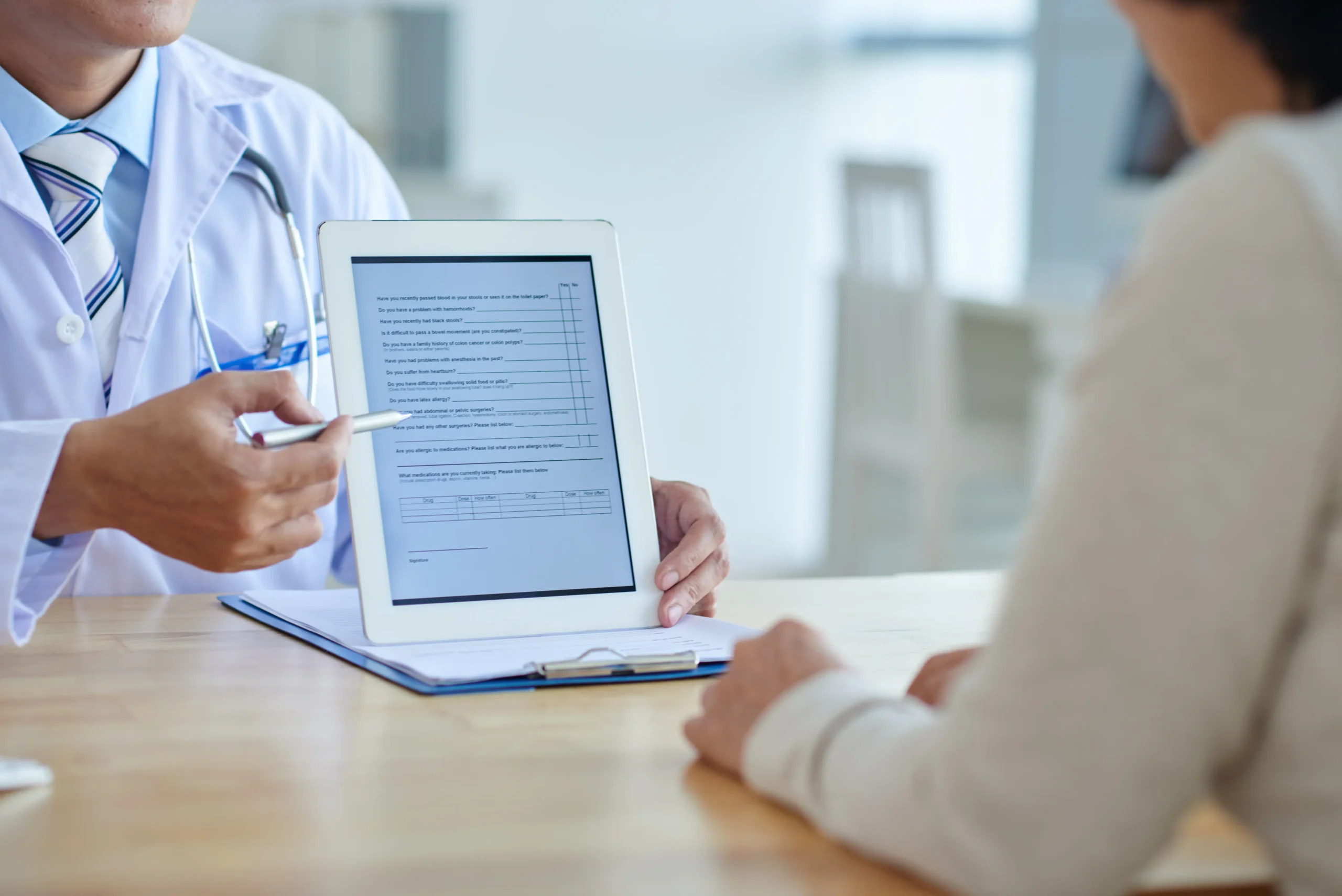 A doctor in a white coat is sitting at a desk with a patient, holding and pointing to a tablet displaying a medical form. The patient, seen from the back, is listening attentively. Both are seated in a brightly lit office environment provided by Doctor management services.