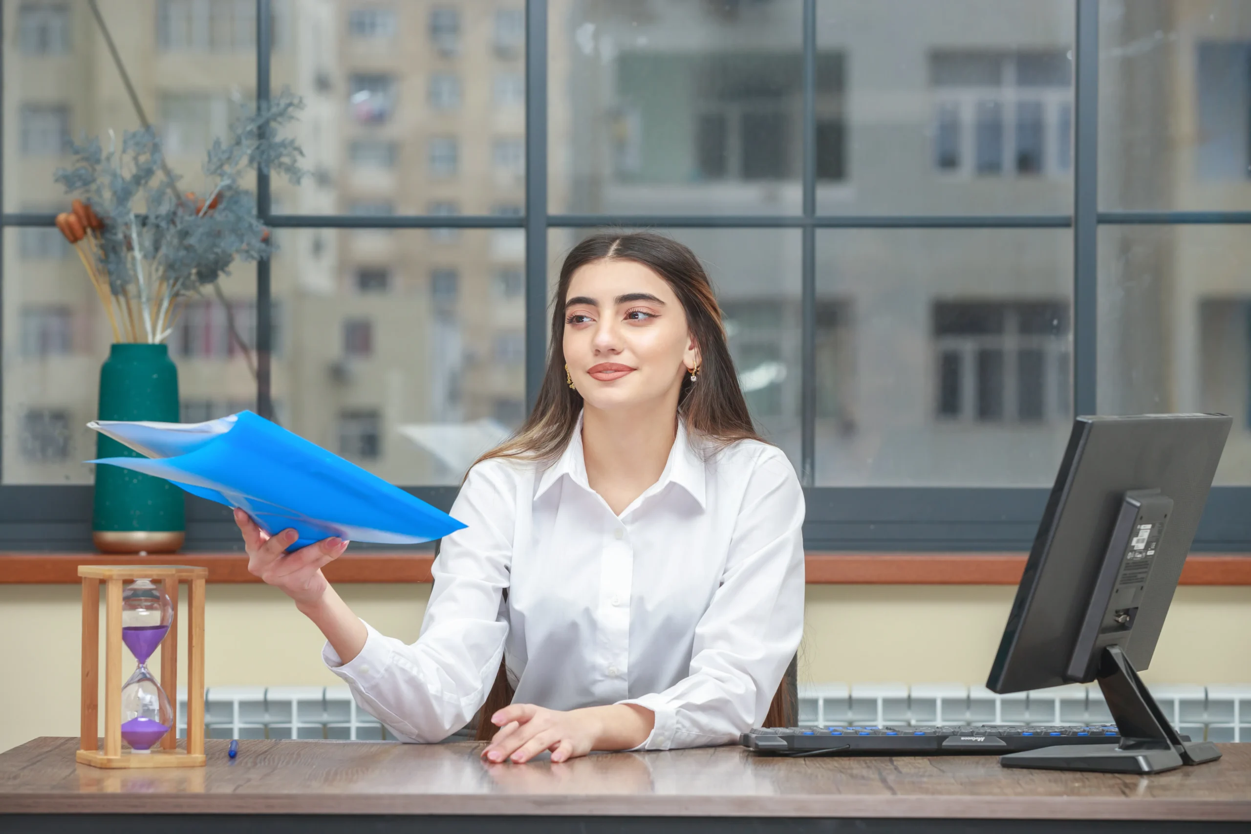 A woman with long brown hair, dressed in a white shirt, sits at a desk holding a stack of blue folders. Behind her is a large window with a cityscape view. On the desk are a computer monitor, an hourglass, and a vase with dried flowers. She appears to be involved in medical billing and coding tasks.