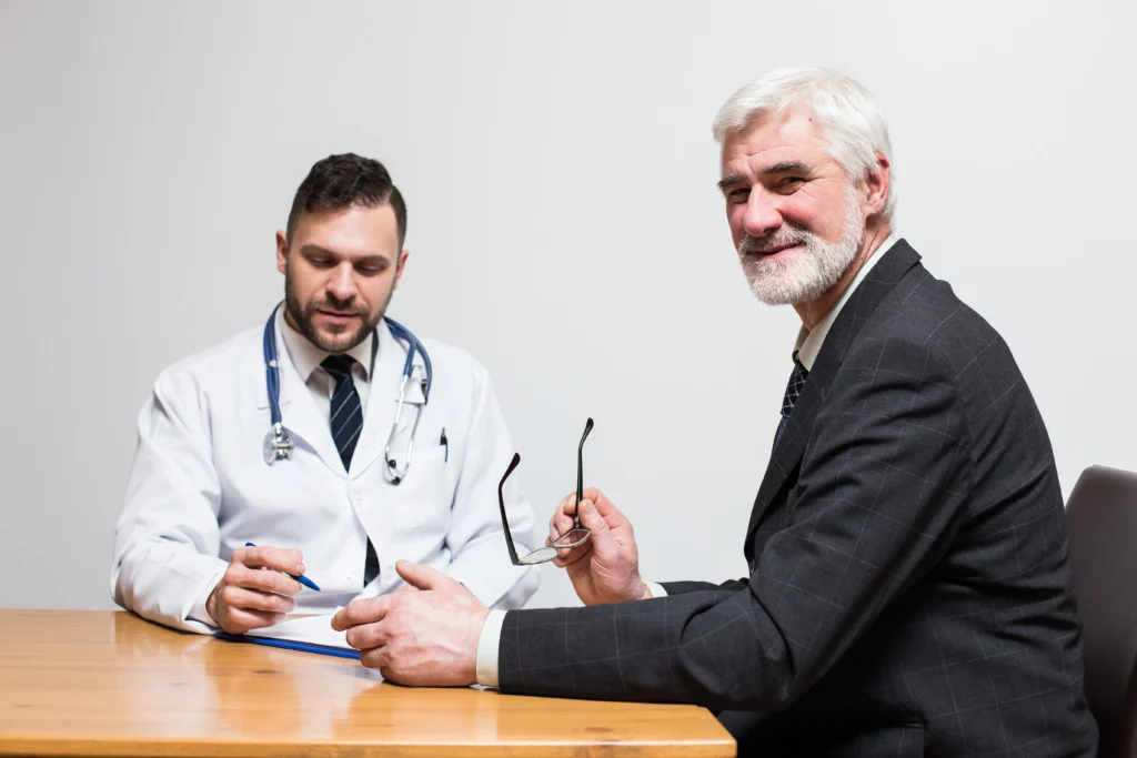 An elderly man with white hair and a beard, dressed in a suit, sits at a table with a younger male doctor wearing a white coat and stethoscope. They appear to be in discussion about doctor management services, with the doctor holding a pen and clipboard and the elderly man holding a pair of glasses.