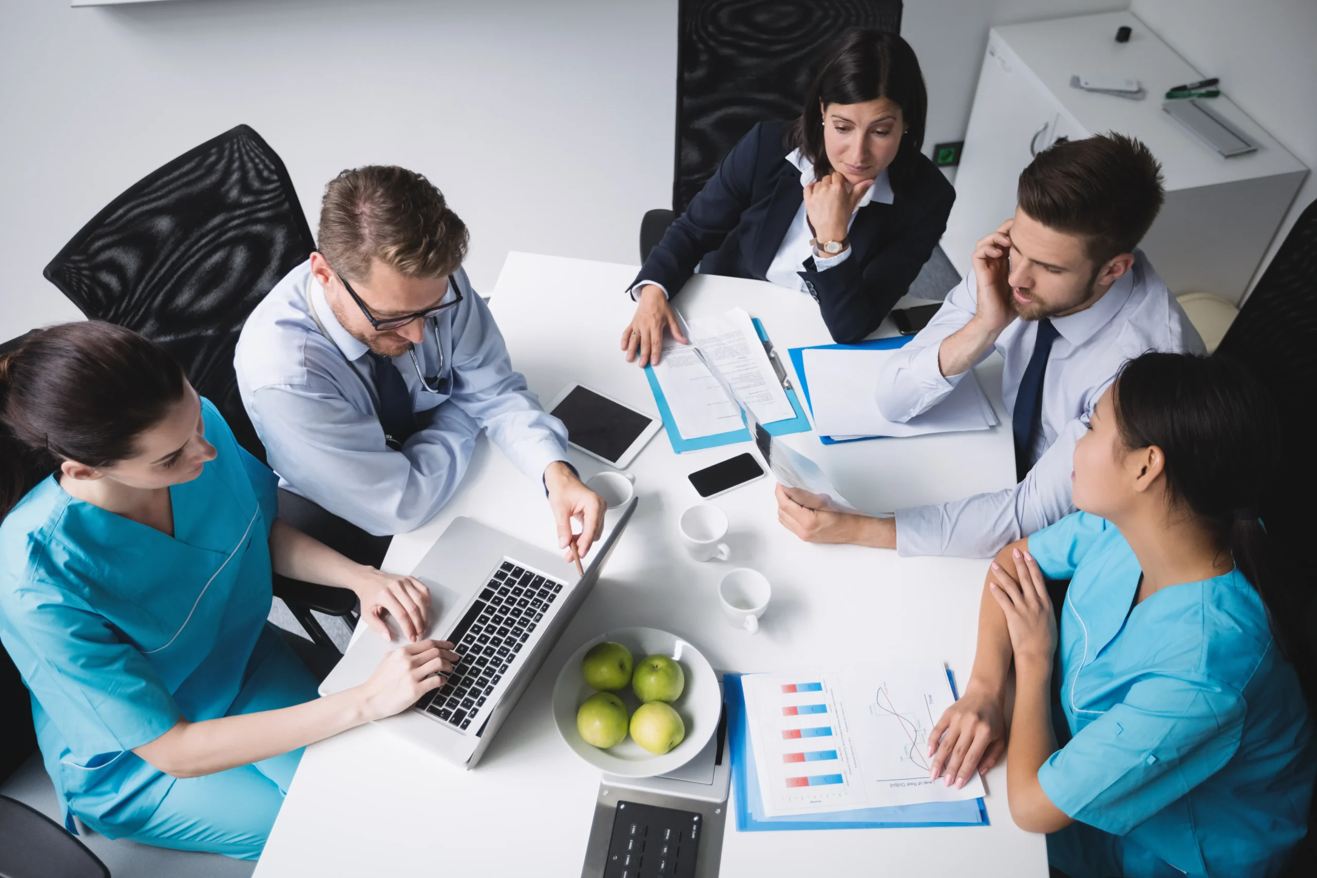 A group of healthcare professionals and business professionals sit around a table. Two people wear medical scrubs, while the others are in business attire. They are using laptops, reviewing documents related to medical billing and coding, and discussing. A bowl of green apples is on the table.