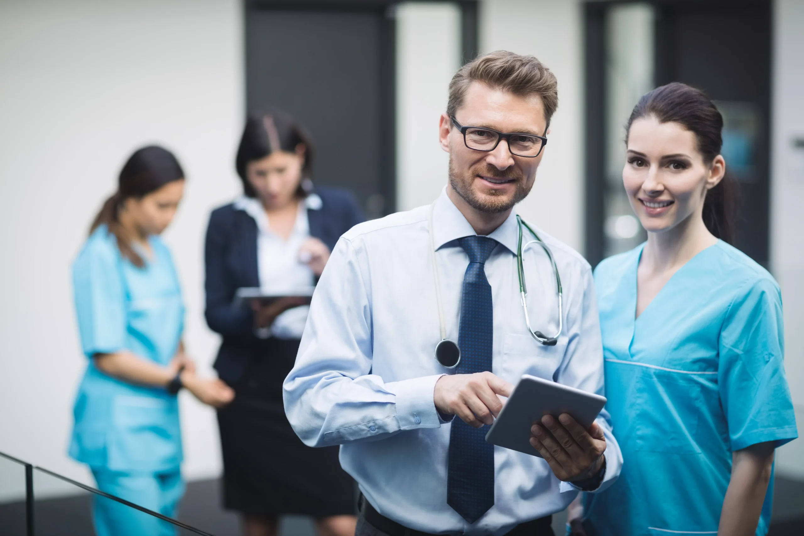 A smiling male doctor, wearing glasses and a stethoscope around his neck, stands next to a female nurse in blue scrubs. Both look at a tablet. In the background, another nurse and a woman in a dark suit discuss documents related to Doctor MGT services.