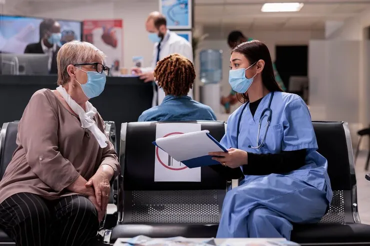 In a hospital waiting area, a healthcare professional in scrubs and a face mask sits next to an older woman with glasses, also wearing a face mask. They appear to be discussing medical billing and coding from a clipboard. Other people and staff are visible in the background.