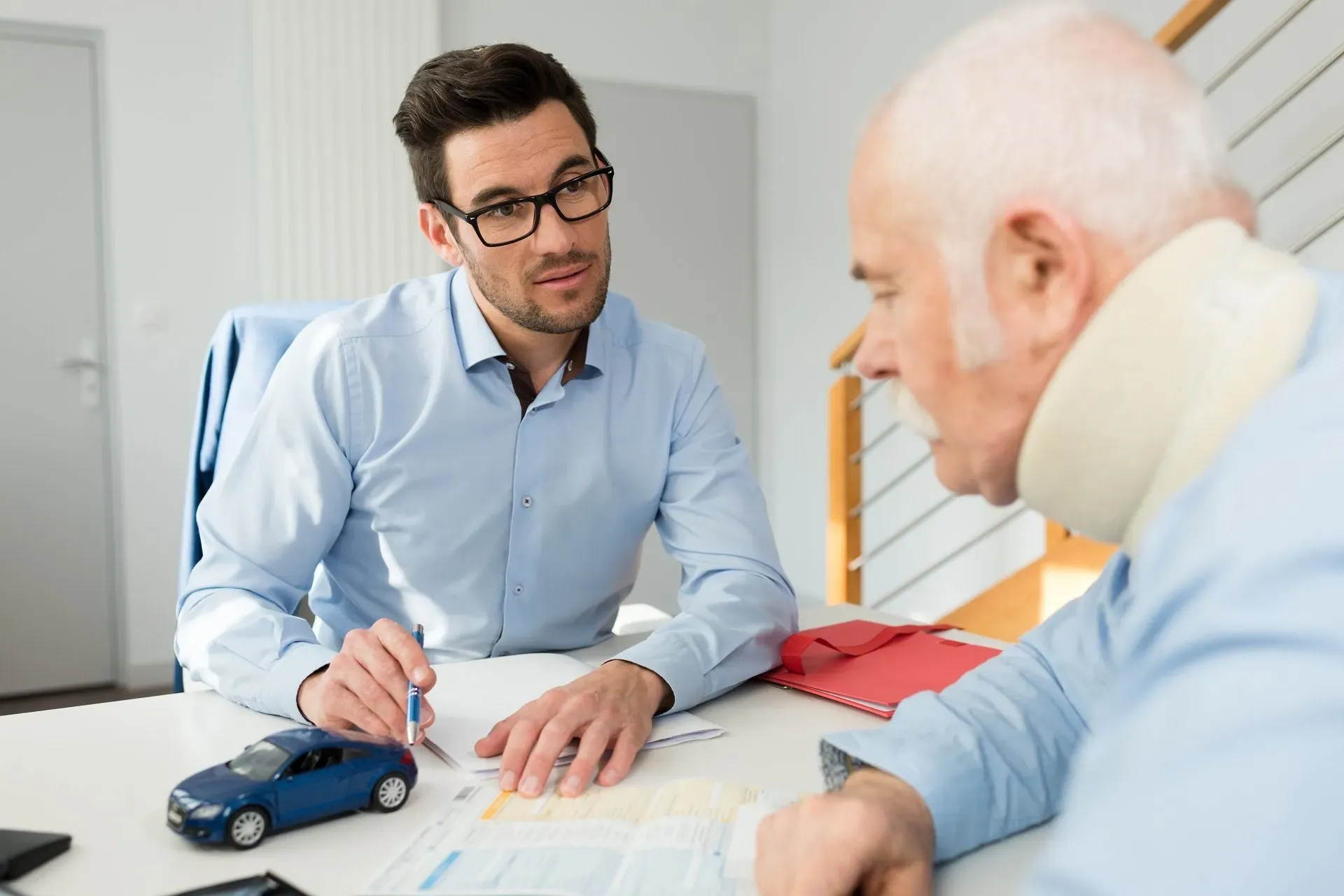 A young man in glasses and a light blue shirt sits at a desk, attentively listening to an older man with a neck brace. The young man holds a pen and looks at documents on the desk, which also has red folders and a small toy car with the Doctor MGT logo.