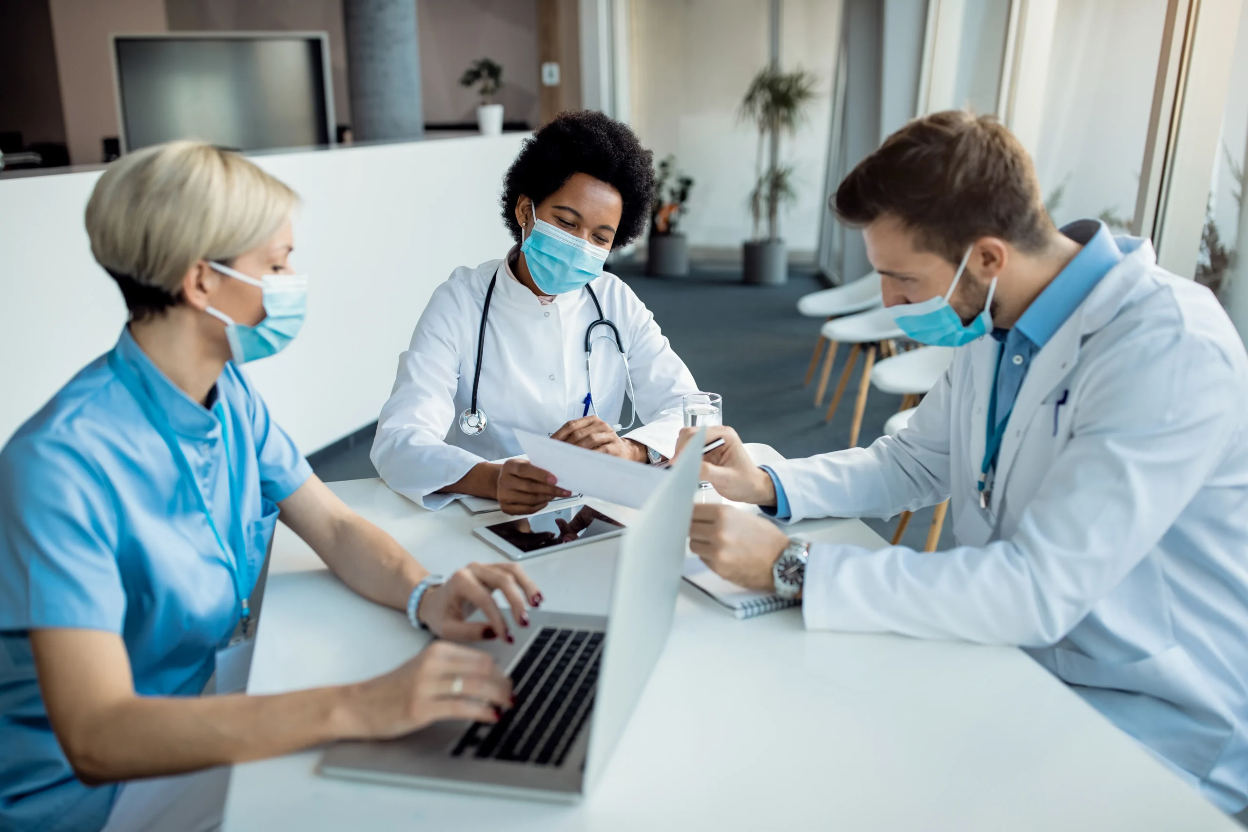 Three medical professionals, wearing face masks, discuss documents at a table in a modern office. One uses a laptop for medical billing and coding, and another writes on a clipboard. They appear engaged and focused on their work, collaborating in a bright, well-lit environment.