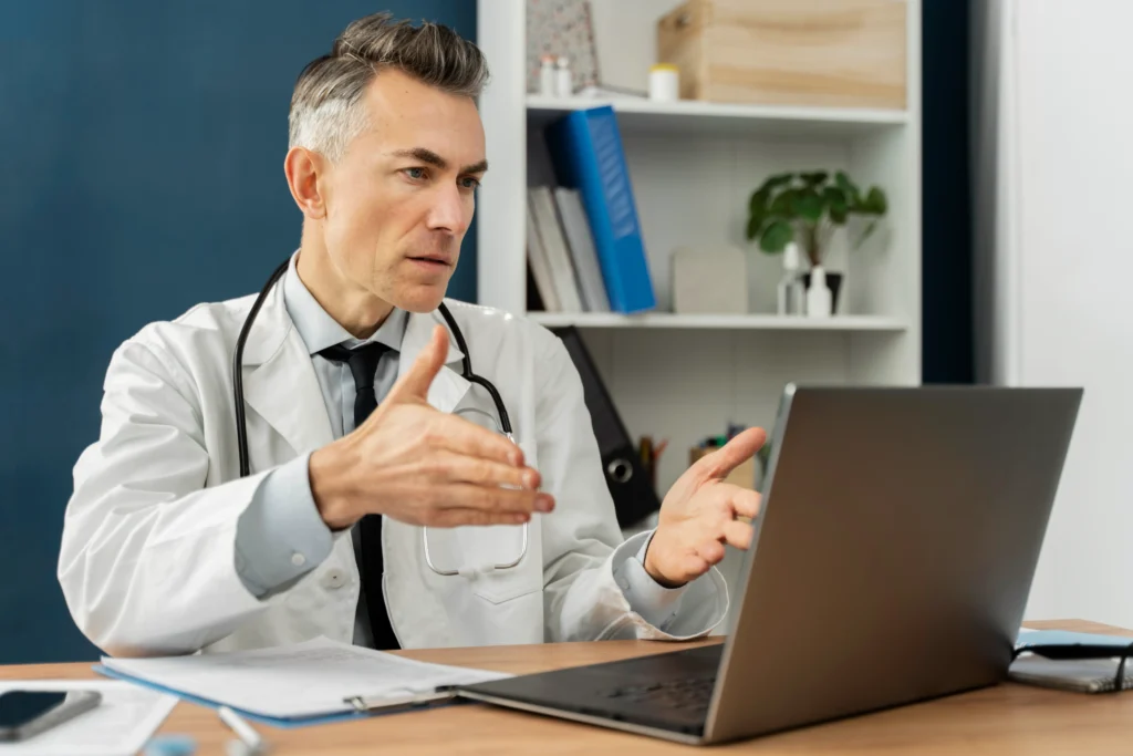 A doctor in a white coat sits at a desk in an office, engaged in a conversation via a laptop. He has a stethoscope around his neck and gestures with his hands while talking. The background includes shelves with books and office supplies, hinting at Doctor management services.