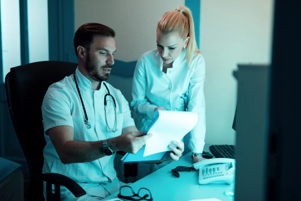 Two medical professionals are examining documents. The man is seated, wearing a white coat and has a stethoscope around his neck. The woman is standing next to him, also in a white coat, intently looking at the papers. They appear to be discussing medical billing and coding in an office or clinic setting.