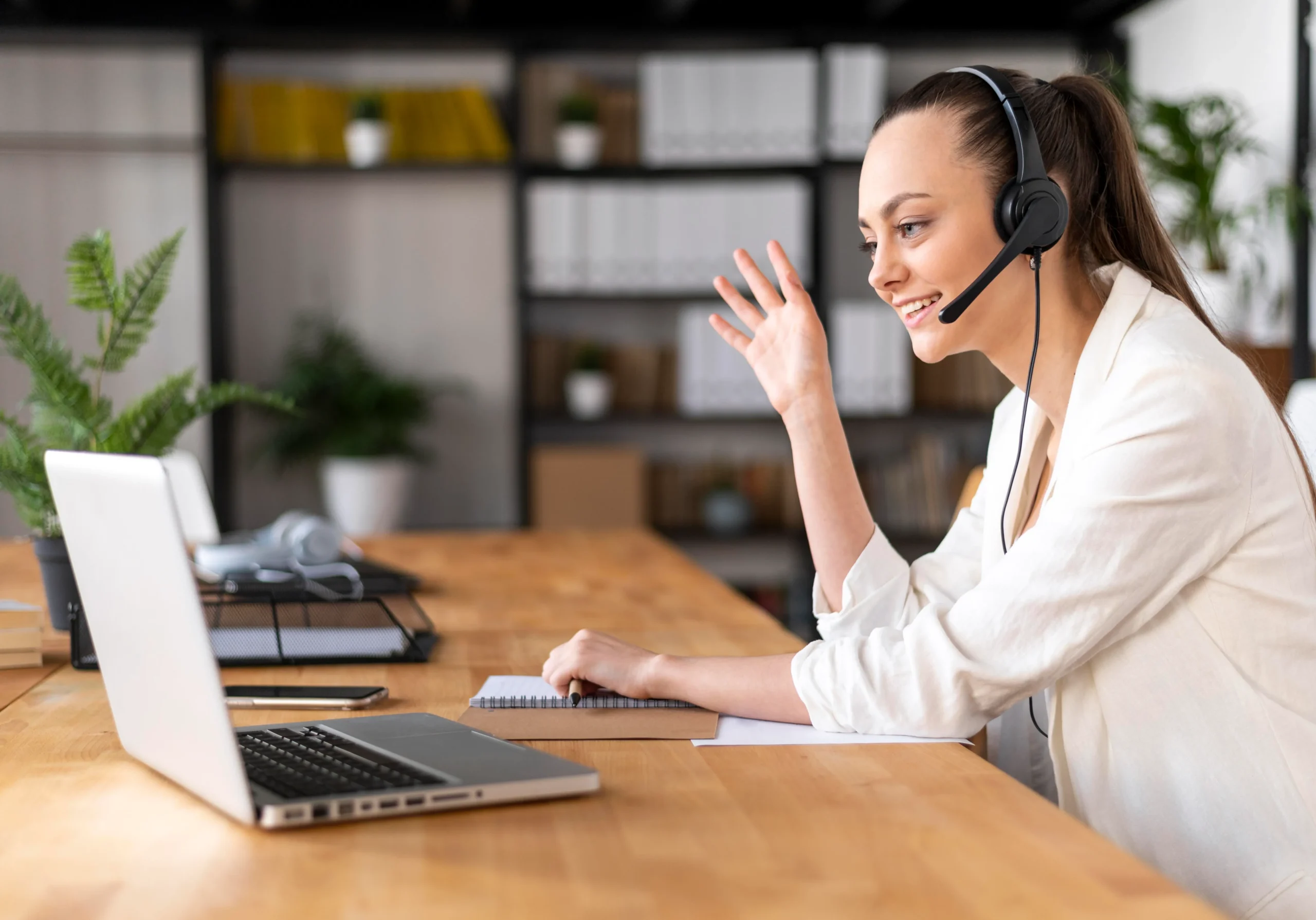 A woman wearing a headset sits at a wooden desk with a laptop, smiling and waving. She appears to be engaged in a video call. The background shows shelves with boxes and plants, creating a tidy and organized workspace, which is likely part of her role in medical billing and coding.