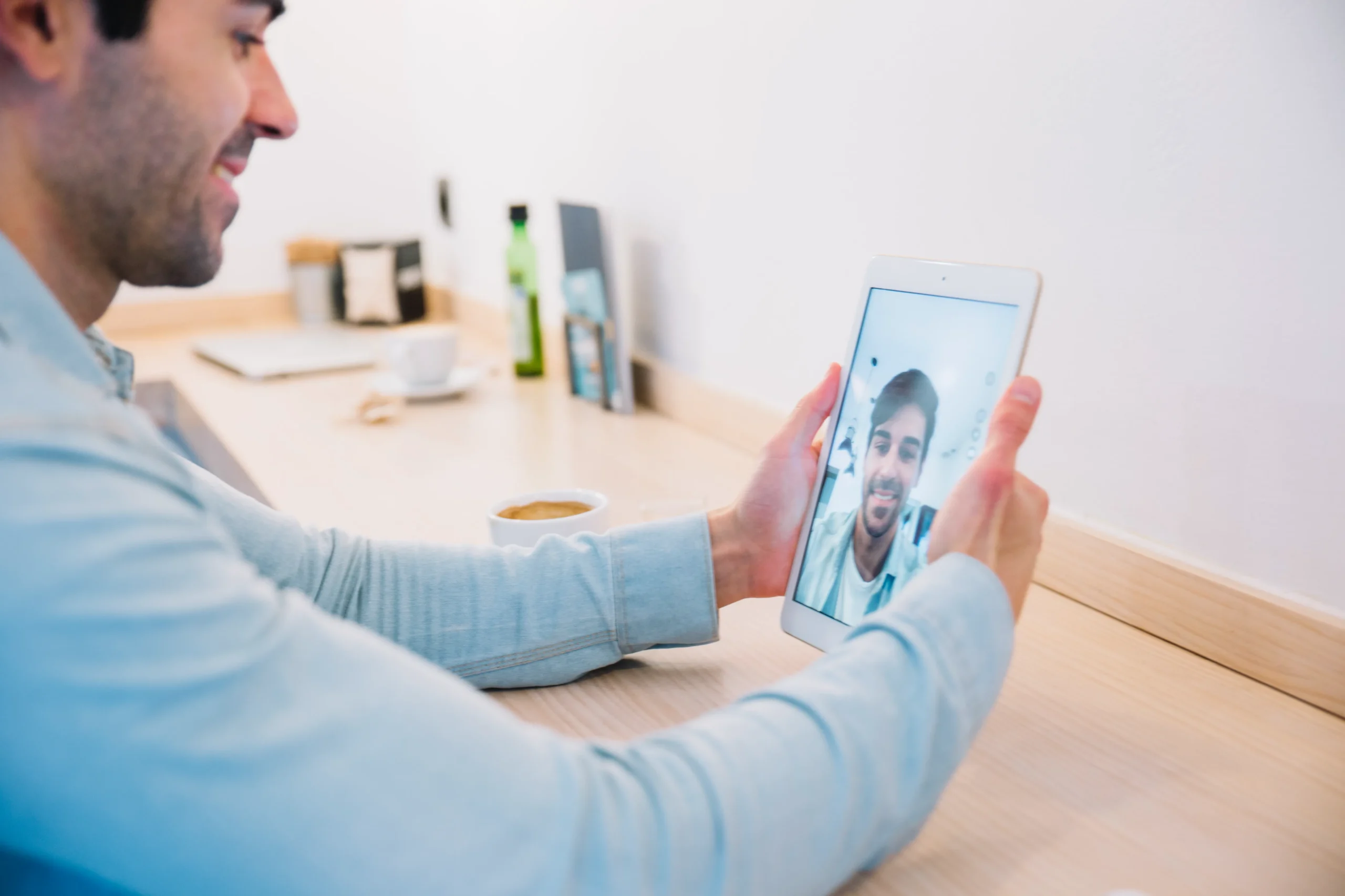 A man sits at a table with a latte, smiling while holding a tablet during a video call. The background features minimalistic decor, including a bottle, a small plant, and a few stationery items related to Doctor MGT.