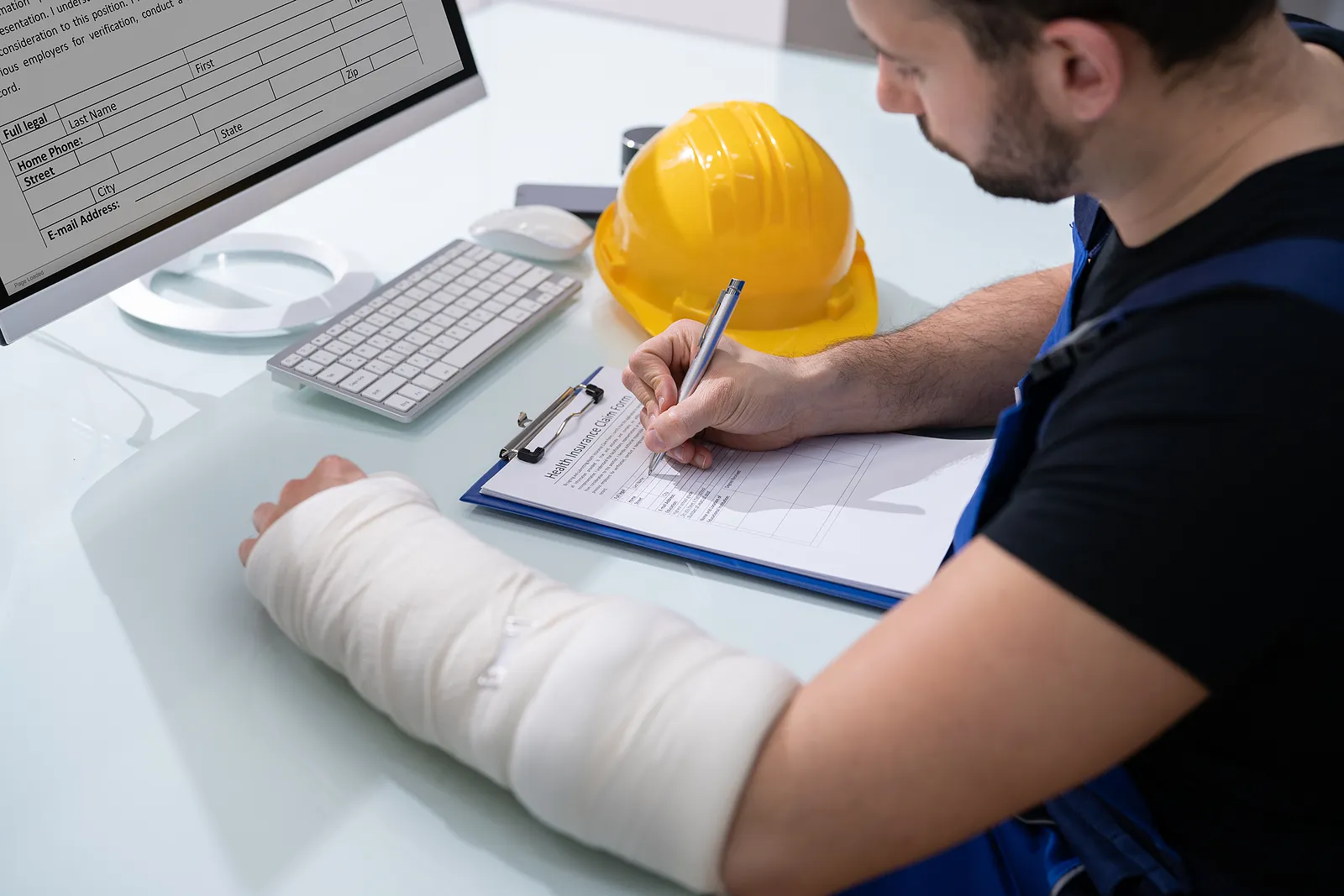 A person with a bandaged arm sits at a desk, filling out a form on a clipboard. A computer monitor, keyboard, mouse, and a yellow safety helmet are also on the desk. The individual appears to be wearing a dark shirt and overalls, possibly involved in medical billing and coding services.