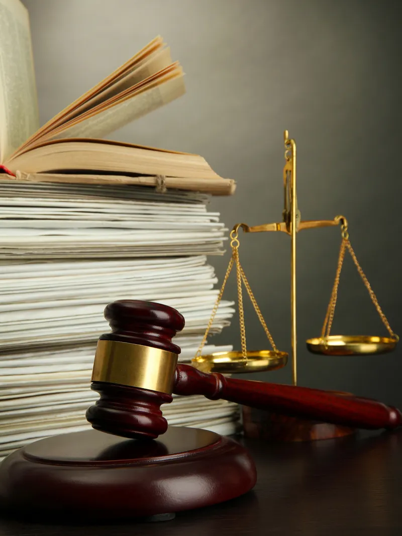 A judge's gavel and sound block sit on a table next to a balanced scale of justice, symbolizing legal fairness. In the background, there is a stack of documents with an open book resting on top. The setting evokes a courtroom or legal office atmosphere relevant to Doctor management services.