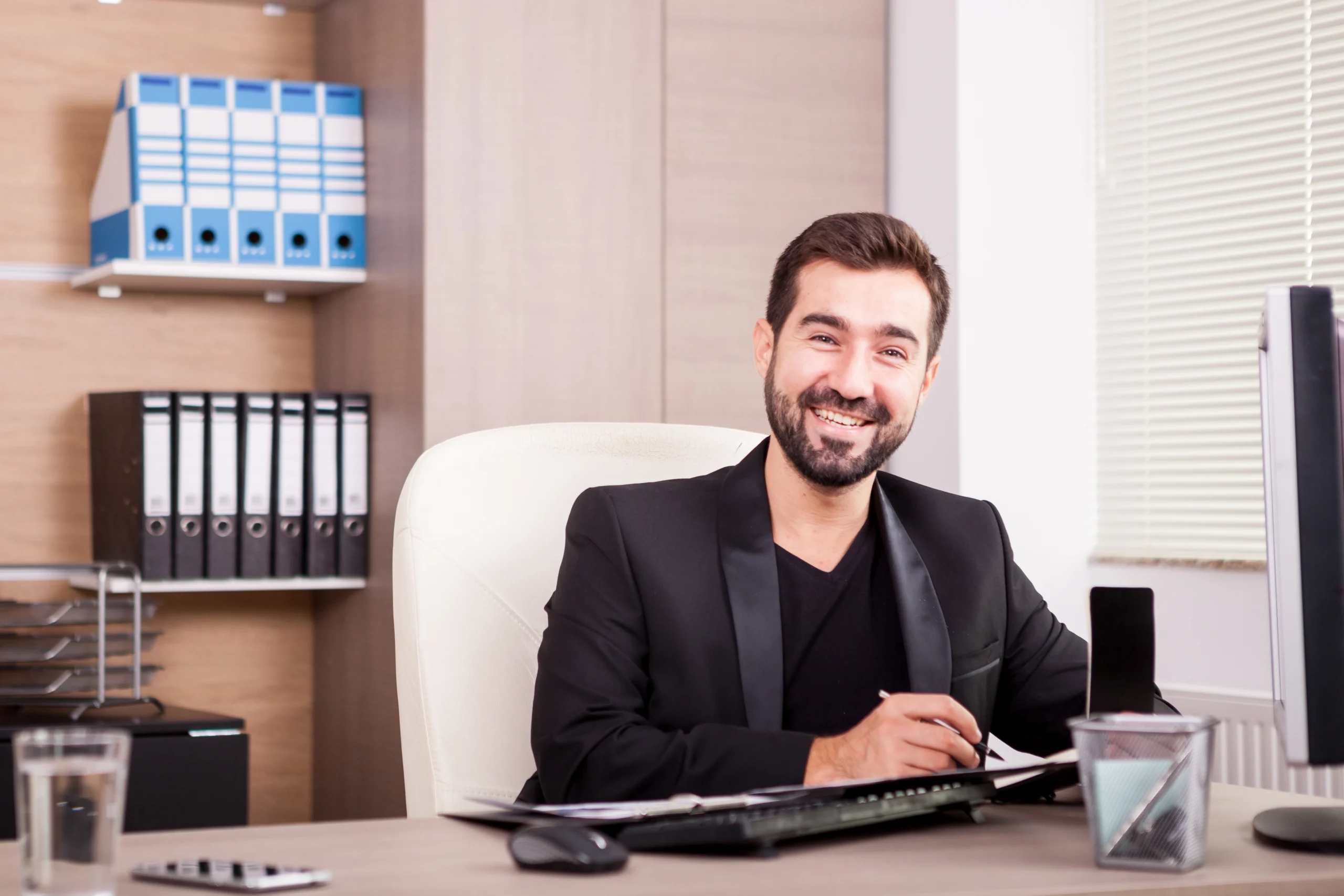 A man with a beard and dark hair is seated at a desk in an office, smiling and wearing a black suit. On the desk are a keyboard, a monitor, papers, and a pen. Shelves with binders and a window with blinds can be seen in the background. The workspace exudes professionalism associated with Doctor MGT services.