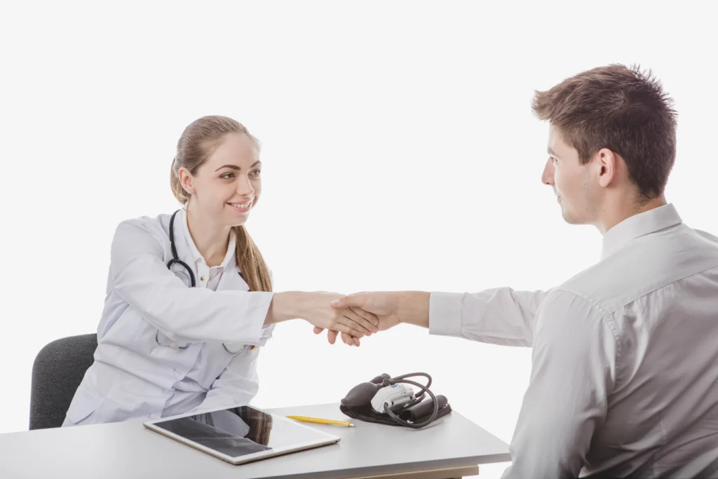 A female doctor with a stethoscope around her neck is shaking hands with a male patient across a desk. The desk has a blood pressure monitor, a tablet, and a yellow pen. Both are smiling, indicating a positive interaction. This scene embodies the professionalism often associated with Doctor MGT services.