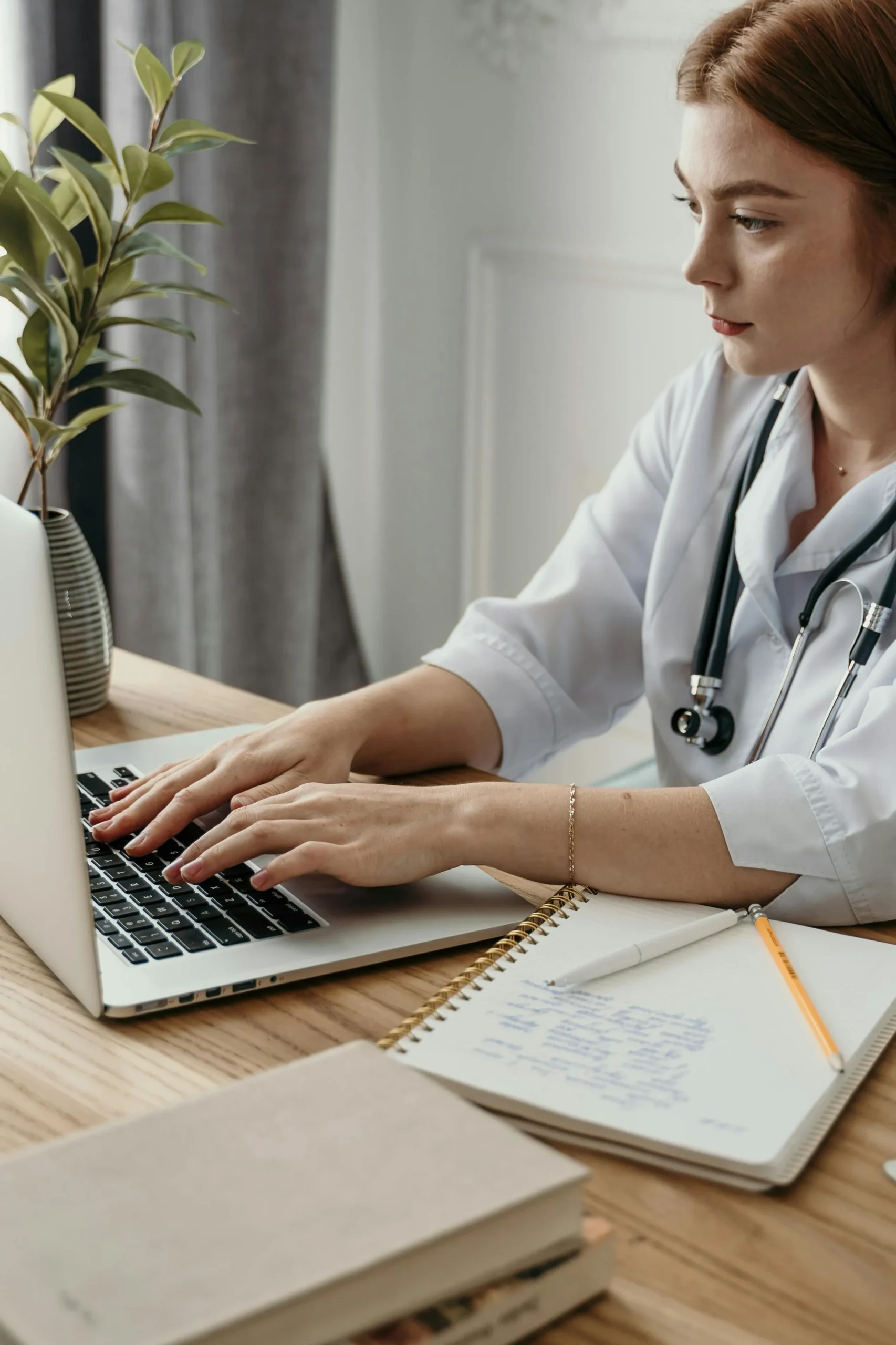 A medical professional with a stethoscope around their neck types on a laptop at a wooden desk, possibly using doctor management services. A notebook with handwritten notes and a pen are placed beside the laptop. A small plant in a vase sits on the desk in the background.